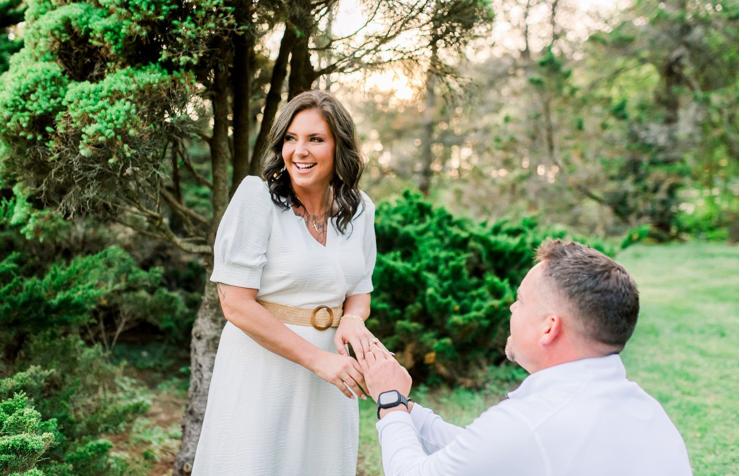 Man on one knee proposing to a smiling woman in a white dress outdoors in a lush green garden.