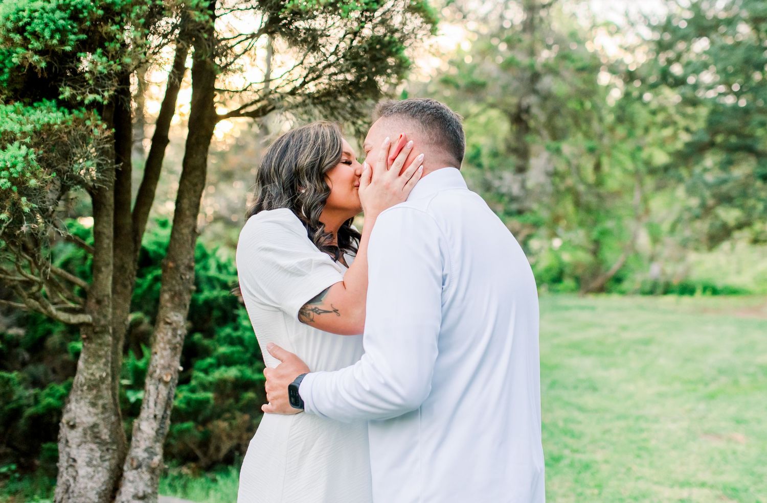 A couple shares a romantic kiss outdoors surrounded by lush green trees in a serene garden setting.