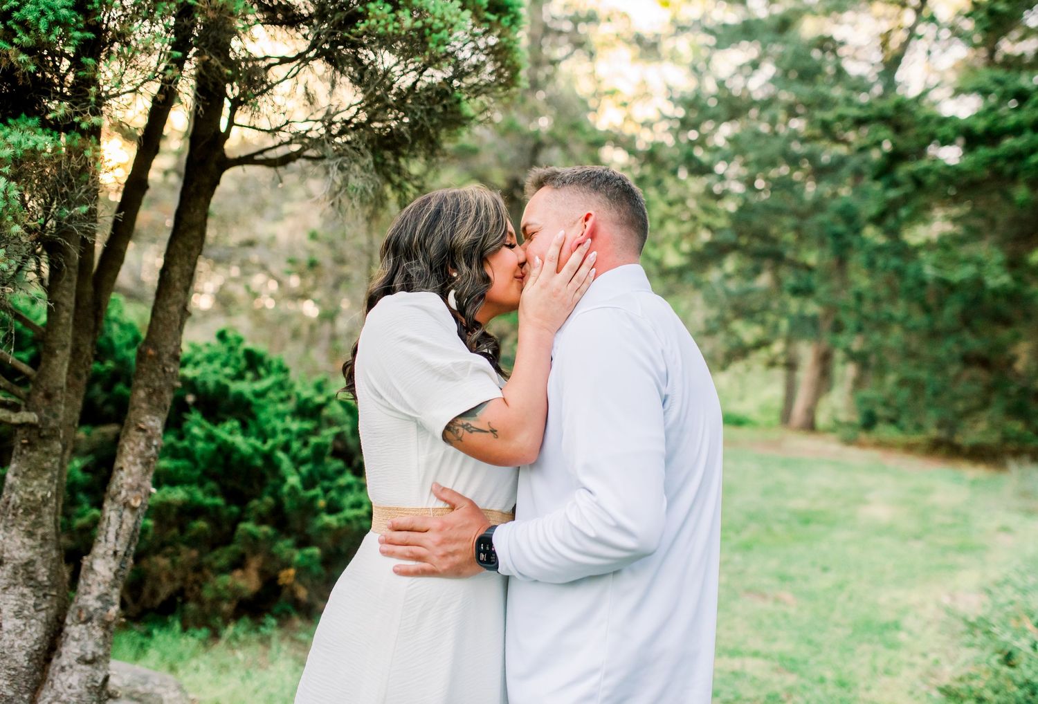 Romantic couple sharing a tender kiss among tall evergreen trees in a sunlit garden setting.