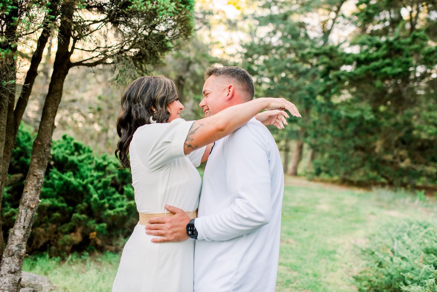 Couple embracing and gazing at each other in a lush green forest setting, both wearing white outfits.