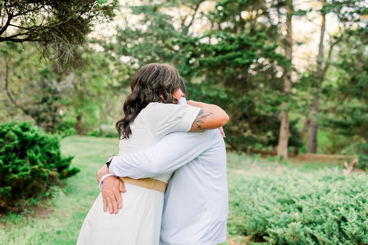 Couple embracing in a romantic hug surrounded by lush green trees during an outdoor engagement session.