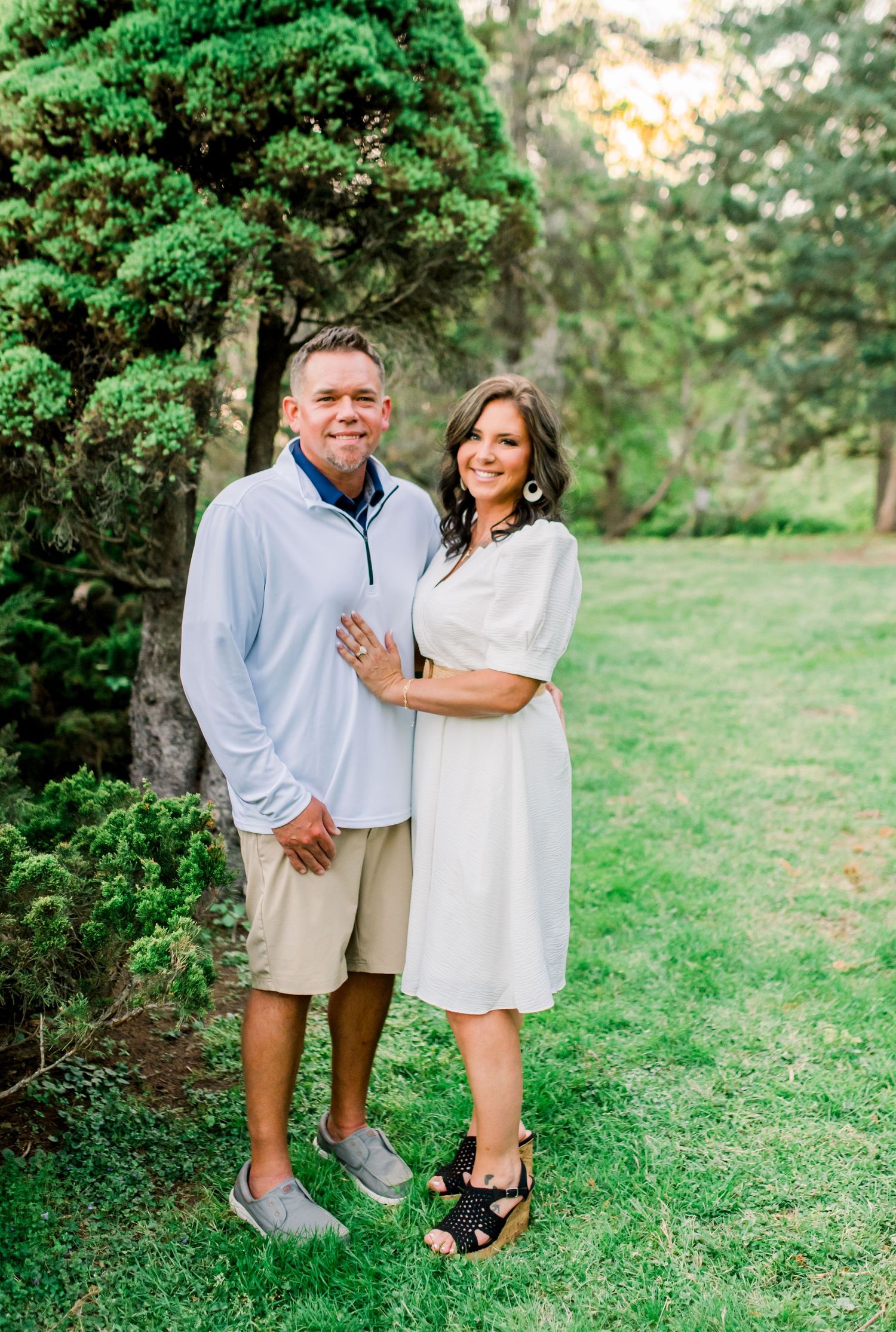 Smiling couple posing together near a decorative evergreen tree, dressed in coordinating casual outfits.