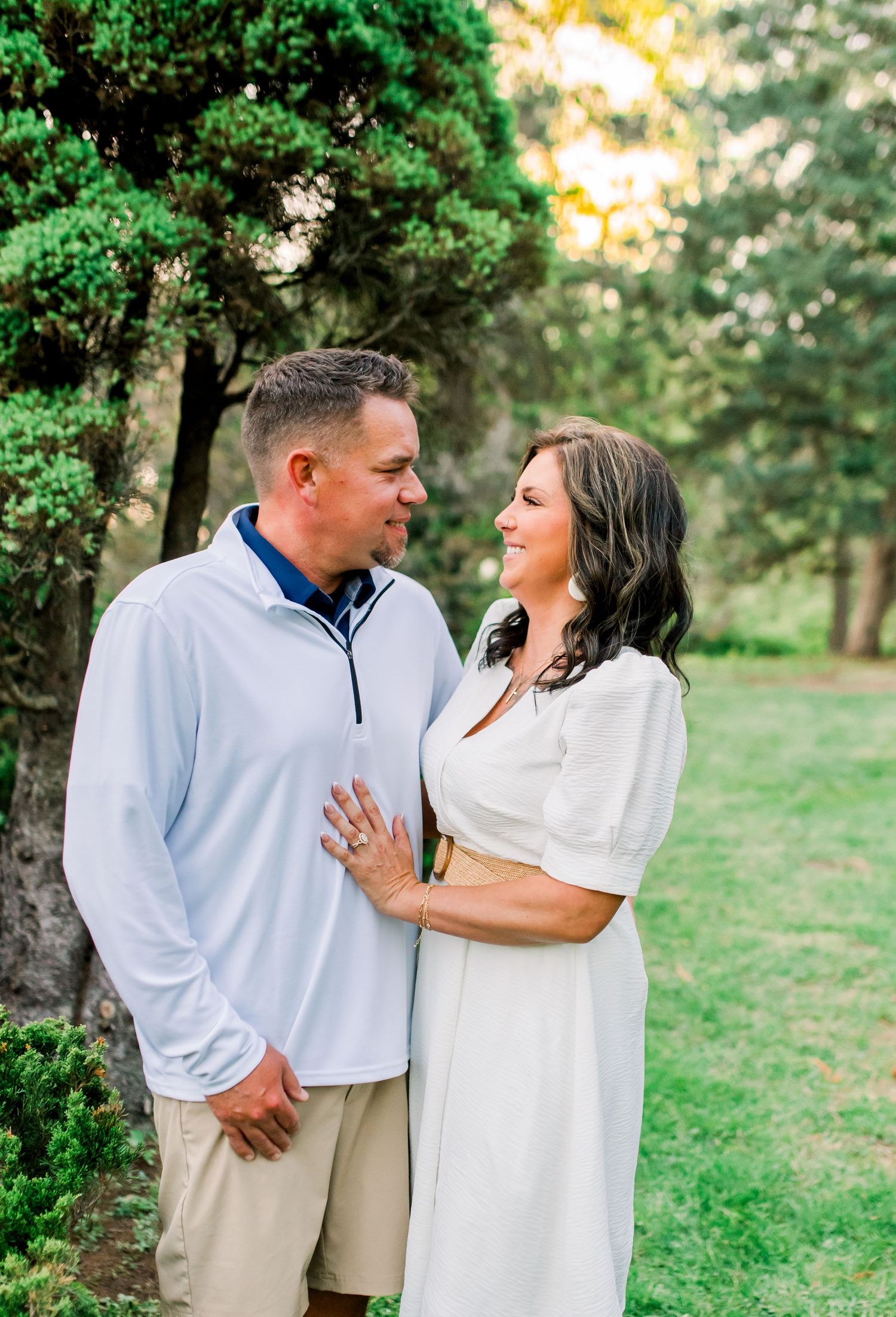 Couple smiling at each other in a lush green park, woman in white dress, man in light blue pullover.