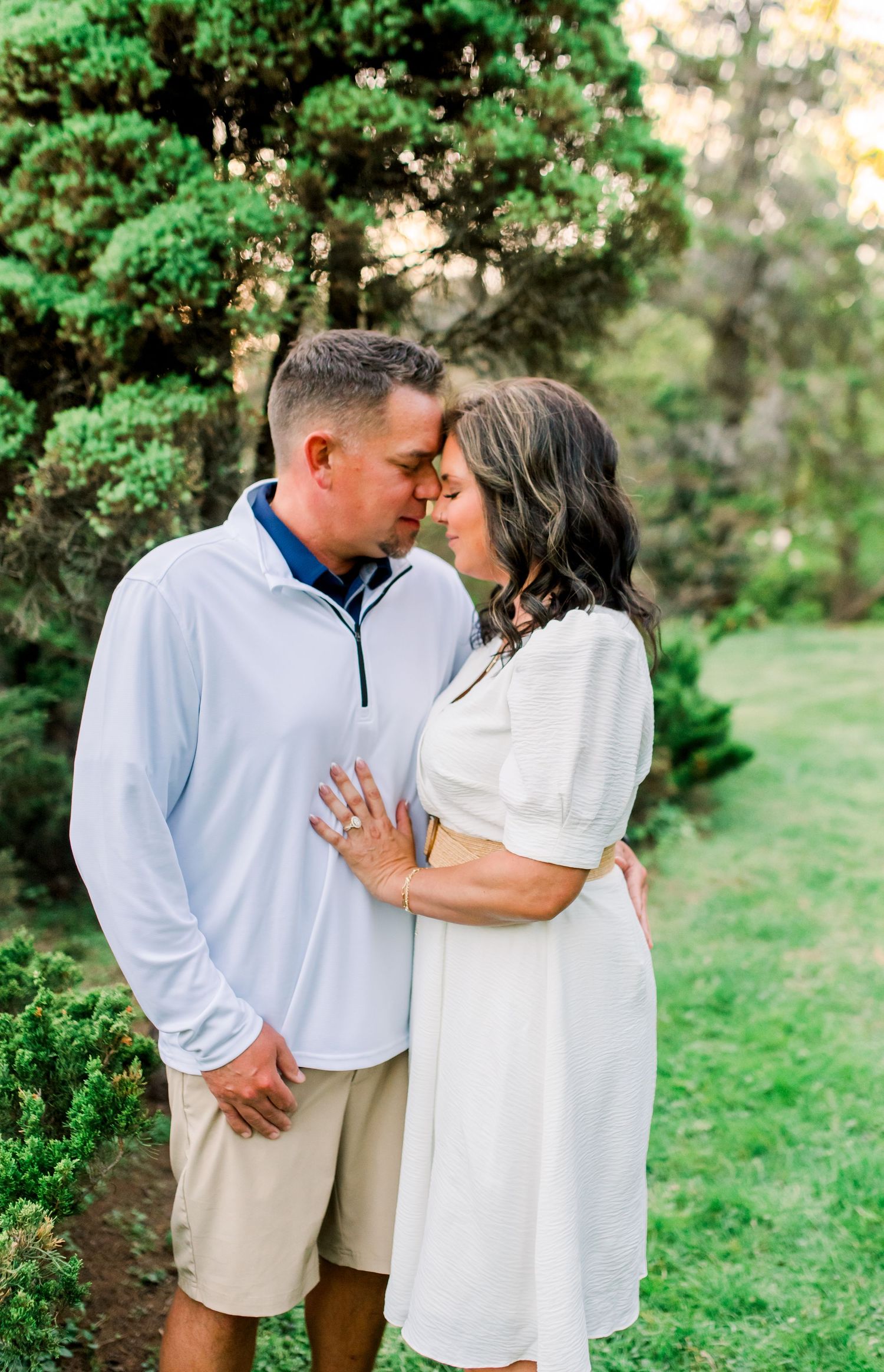 Loving couple in white outfits touching foreheads intimately while standing in a lush green garden.