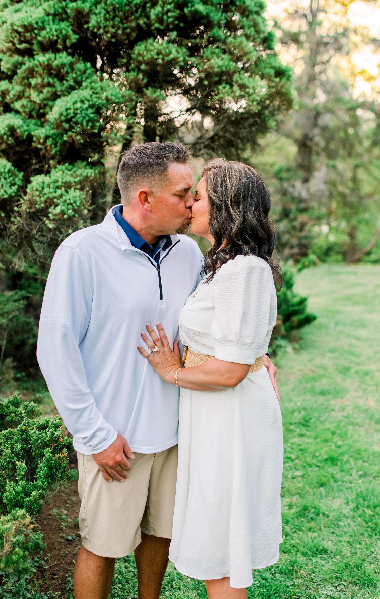 Couple sharing a romantic kiss outdoors near evergreen trees, woman wearing white dress with engagement ring visible.