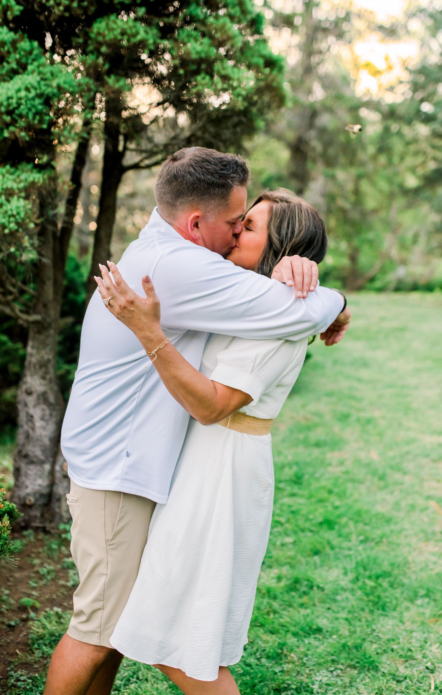 Happy couple embracing and kissing outdoors in a green garden with tall trees in the background.