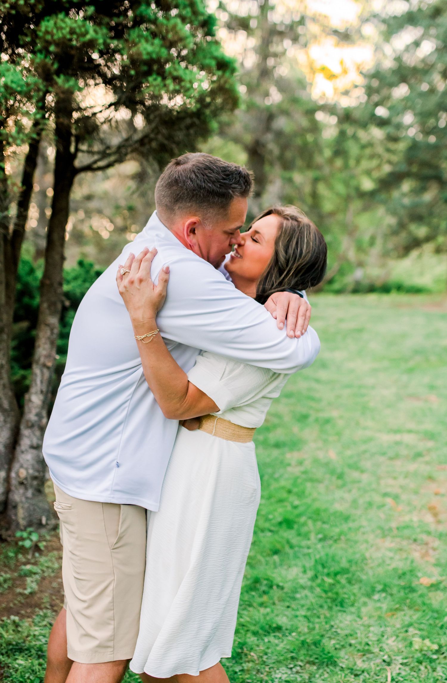 Happy couple embracing and kissing outdoors surrounded by green trees during golden hour.