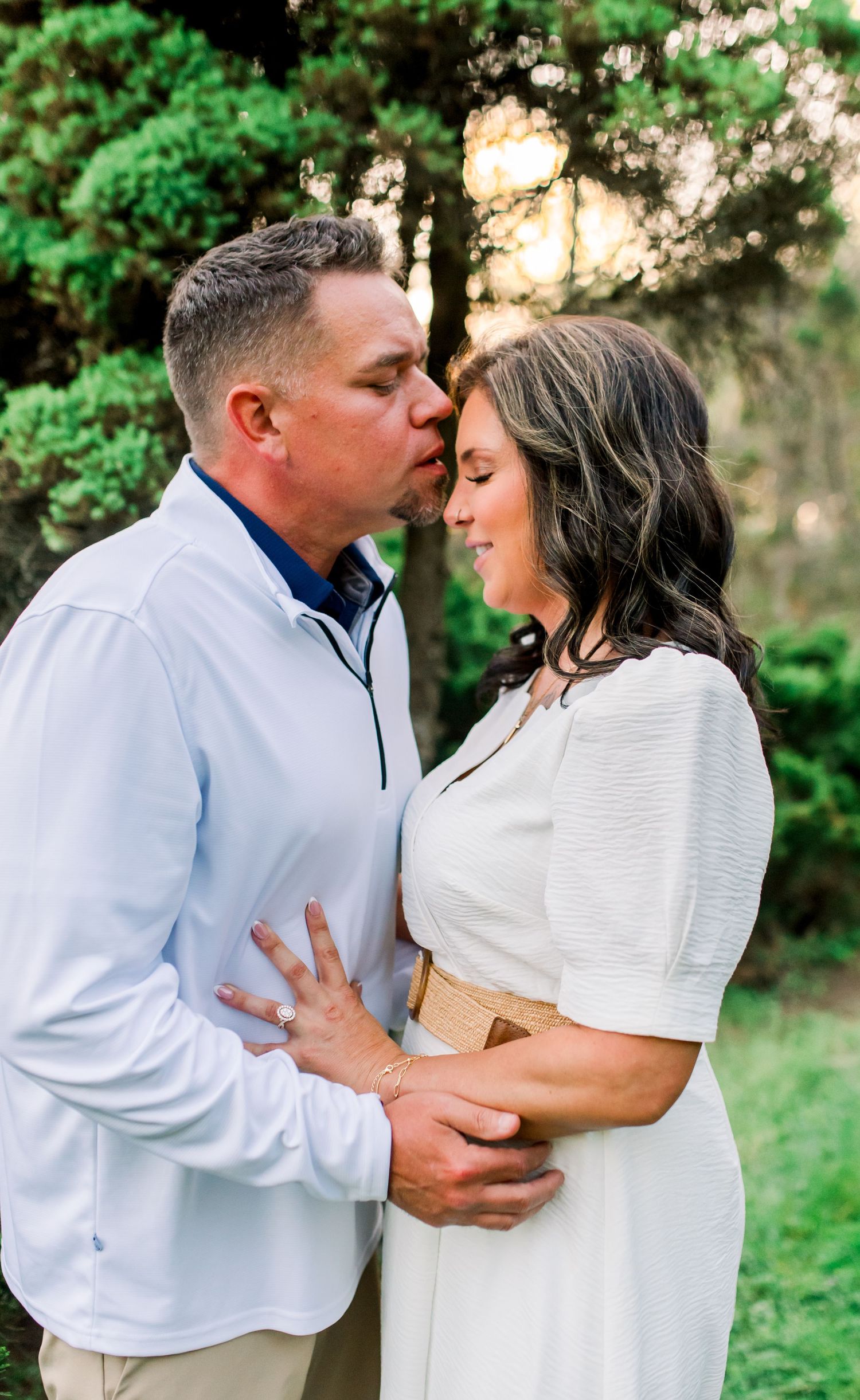 Man in light blue pullover kissing woman on cheek while holding her close in a garden setting.