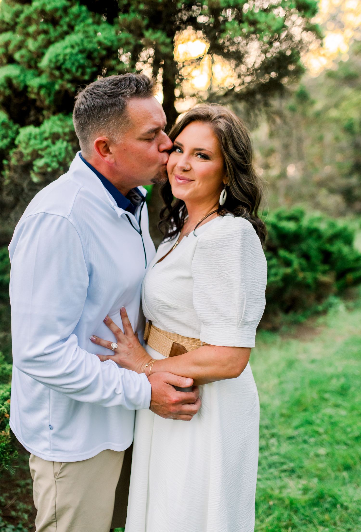 Man in light blue shirt kissing woman in white dress on the cheek in a lush garden setting.