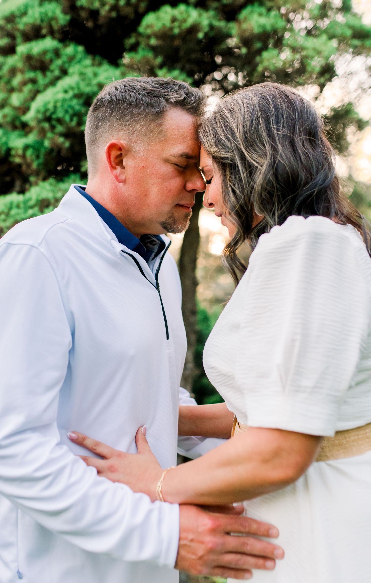 A couple shares an intimate forehead touch outdoors, dressed in white, surrounded by lush green trees.
