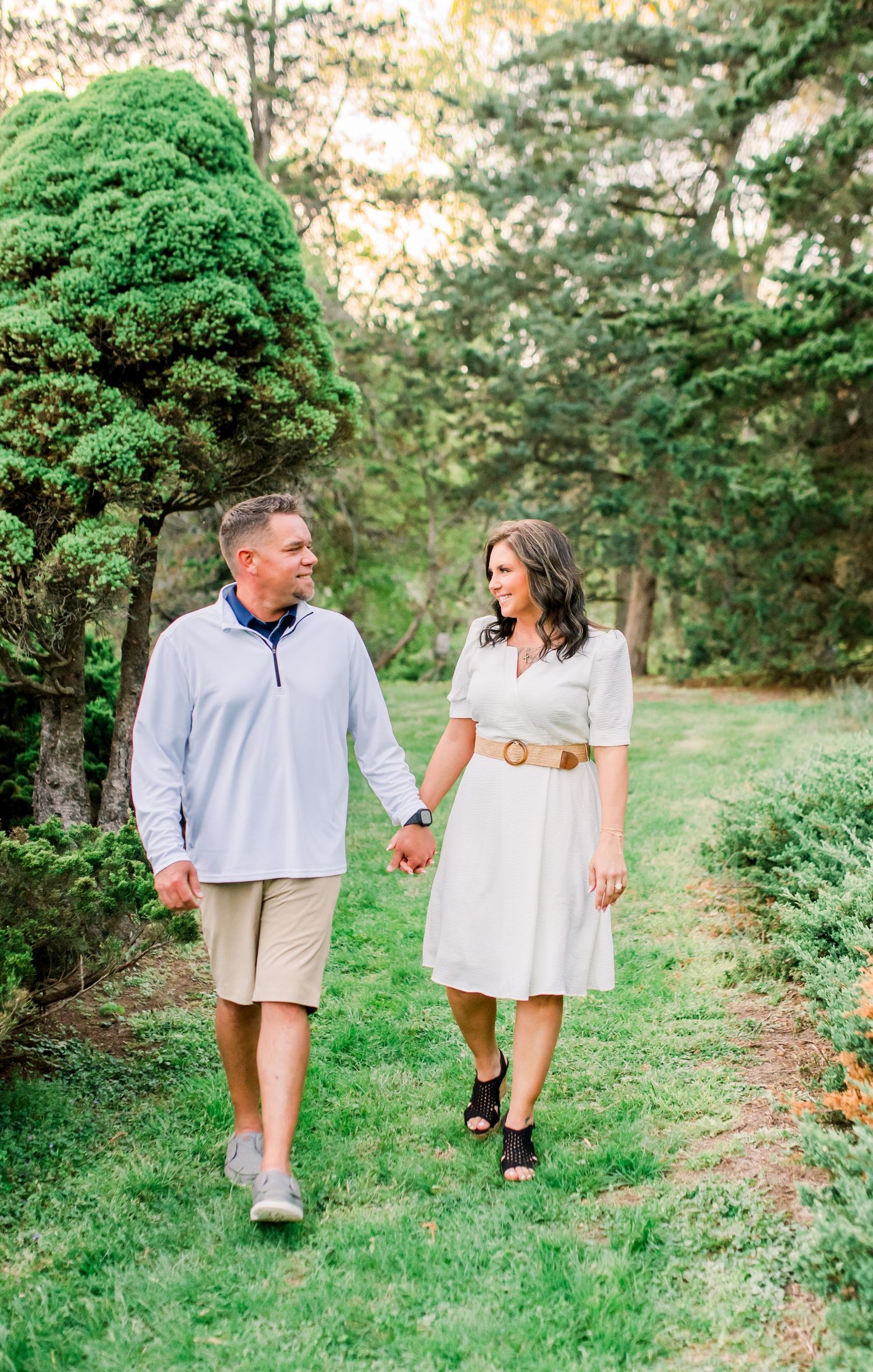A couple walks hand-in-hand through a lush green garden path, smiling at each other on a sunny day.