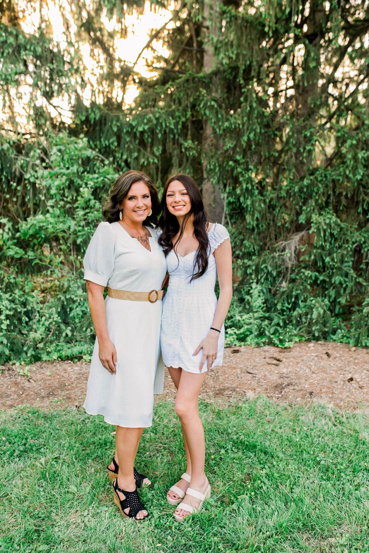 Two women in white dresses smiling together outdoors on green grass with lush evergreen trees behind them.