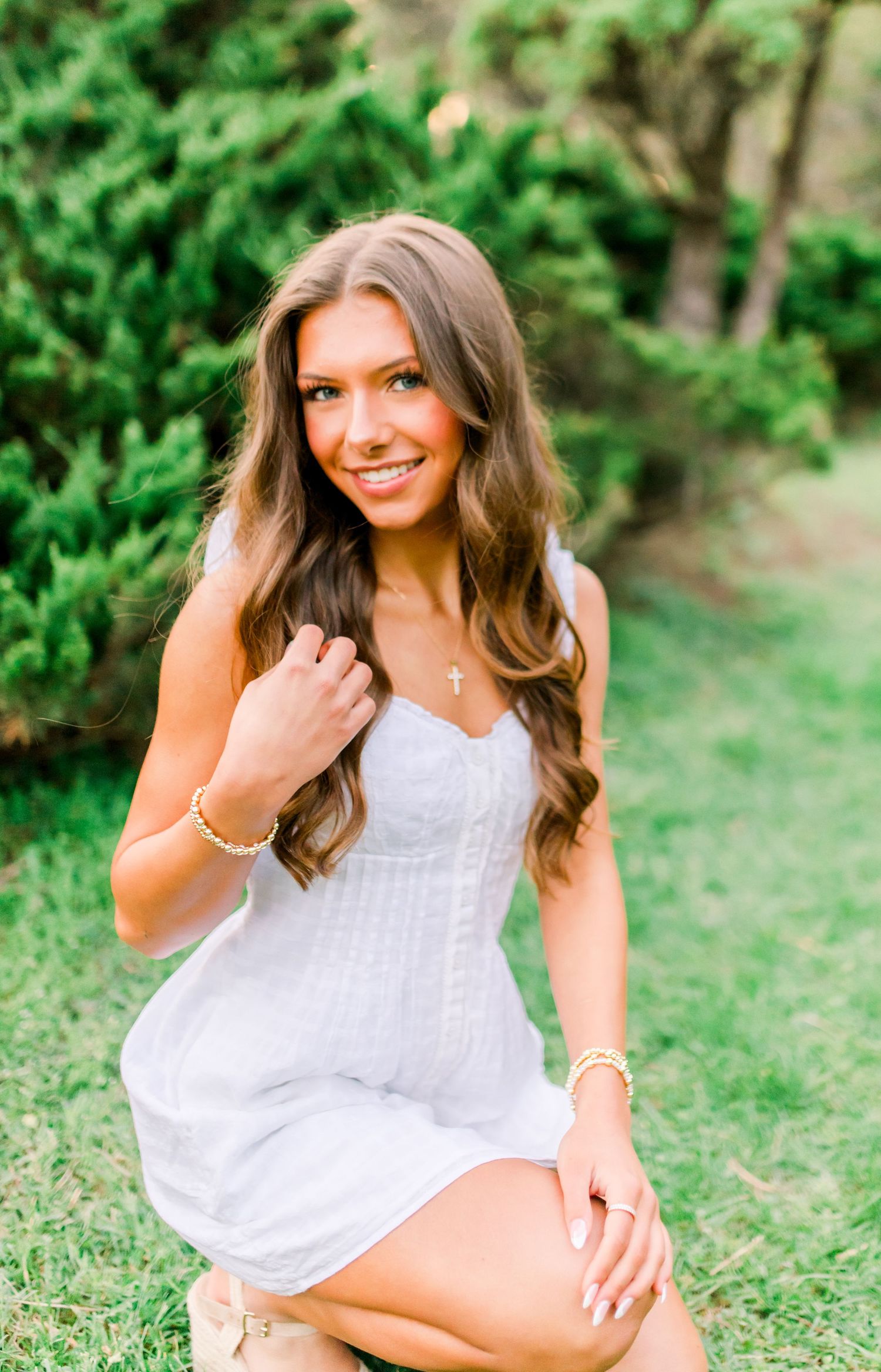Young woman in white dress posing outdoors on green grass with lush trees in background, wearing gold jewelry and cross necklace.