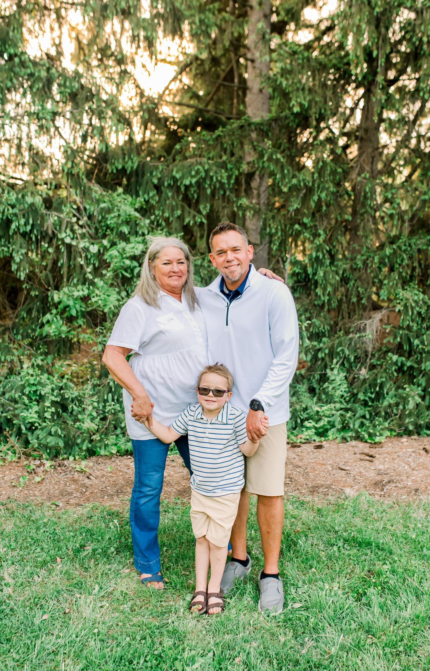 Three family members pose outdoors on green grass with lush trees, including a woman, man, and young child wearing sunglasses.