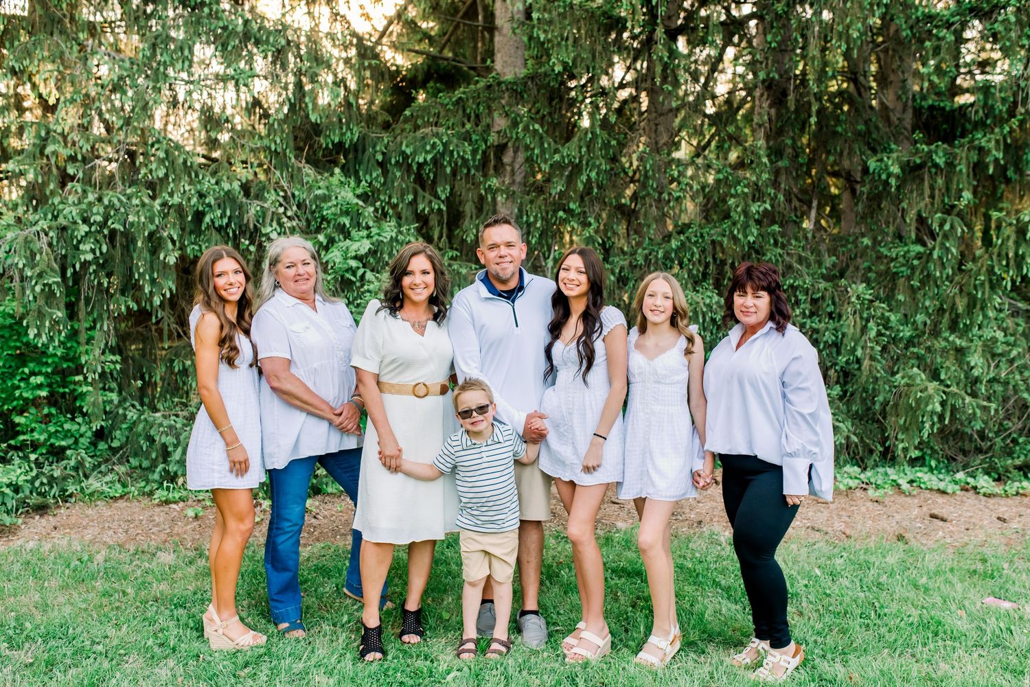A joyful multigenerational family poses together outdoors on green grass with lush trees in the background.