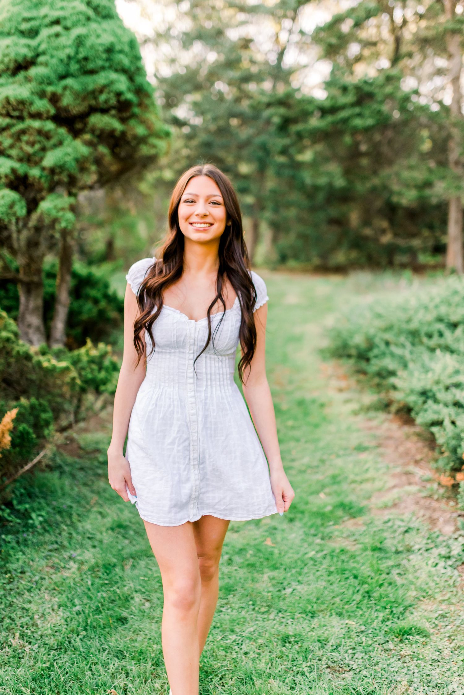 Young woman in white mini dress smiling while walking on a lush green garden path surrounded by trees.