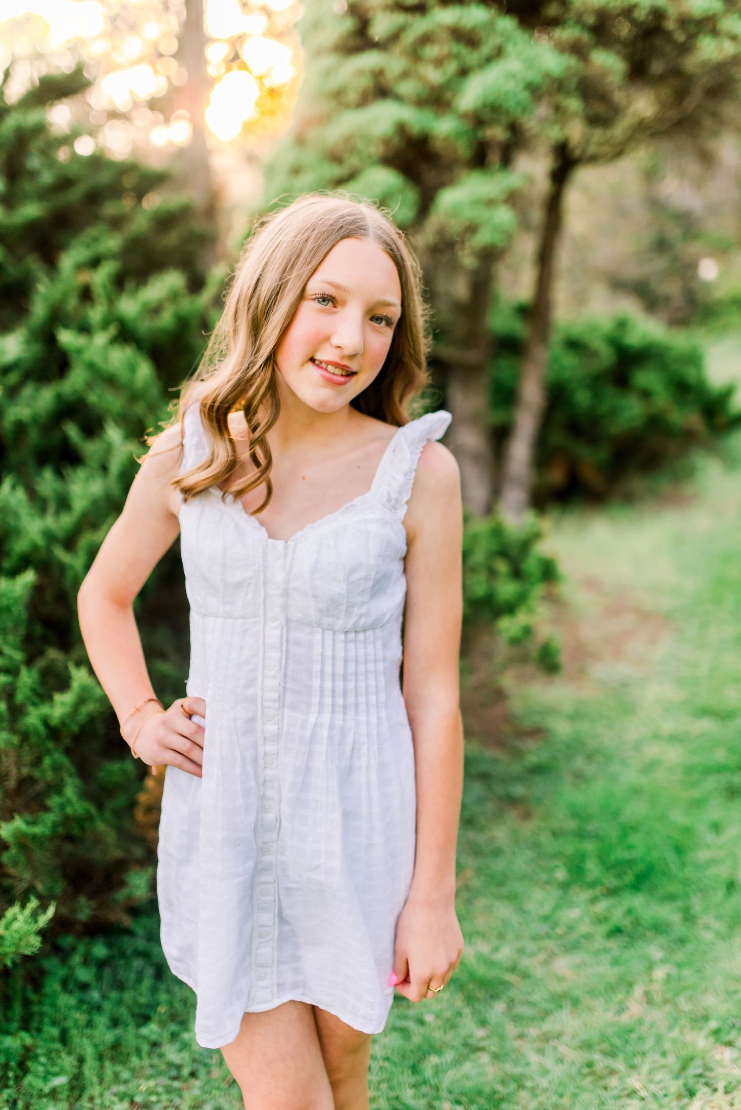 Young girl in white sundress smiling outdoors among green trees in golden hour light.