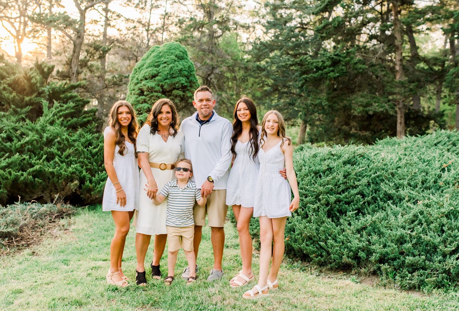 Happy family of six posing outdoors in coordinated neutral outfits surrounded by lush green trees.
