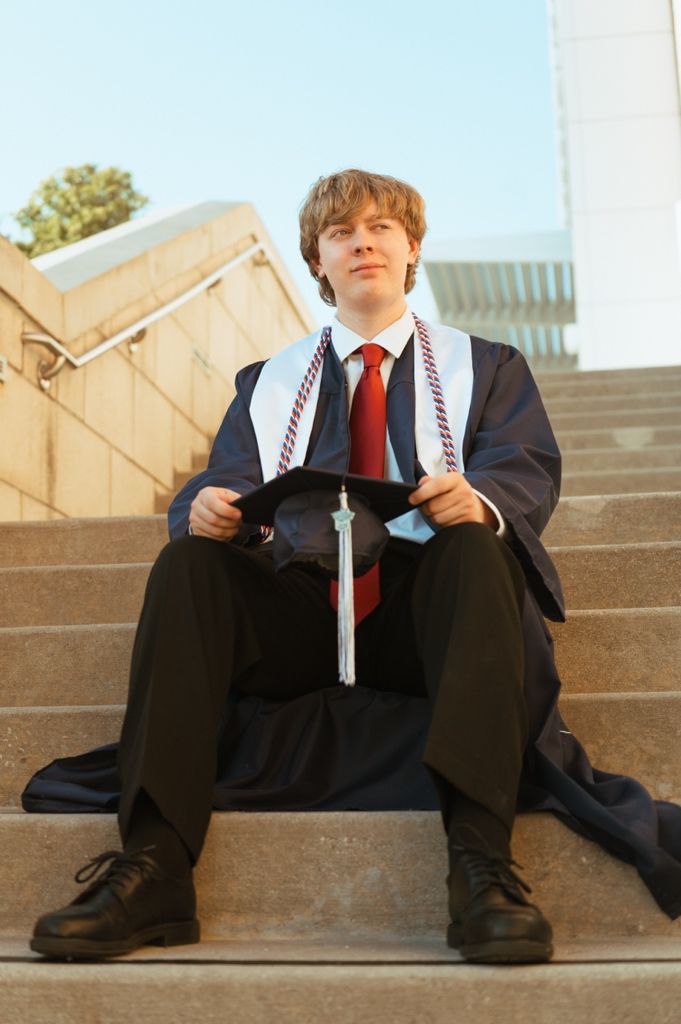 Male graduate in cap and gown sitting on stone steps holding his mortarboard, wearing red tie.