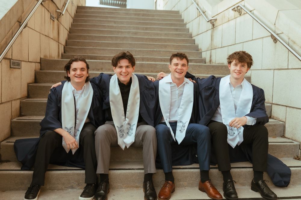 Four young men in graduation attire with stoles smile together on stone steps.