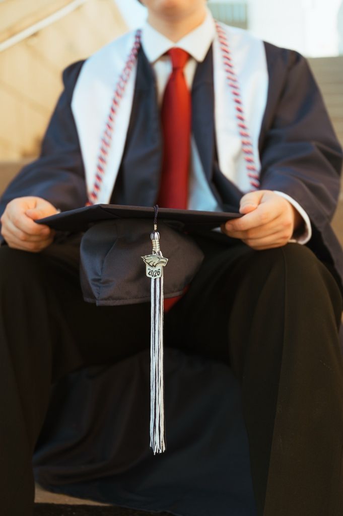 Close-up of graduate in cap and gown holding black mortarboard with silver tassel and honor cords.