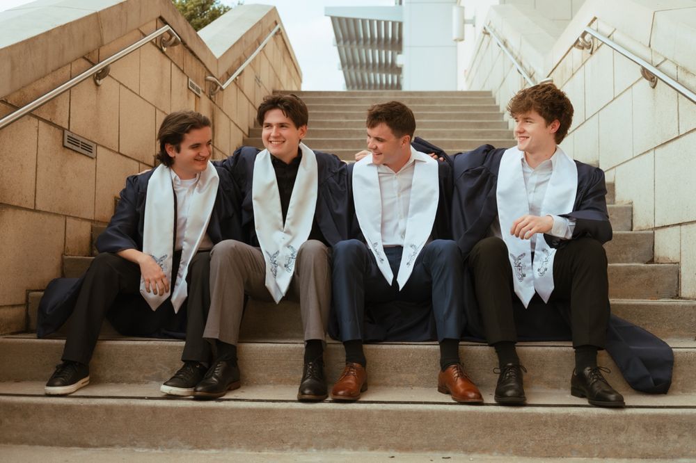 Four male graduates in dark suits and white stoles sit laughing on outdoor stone steps.