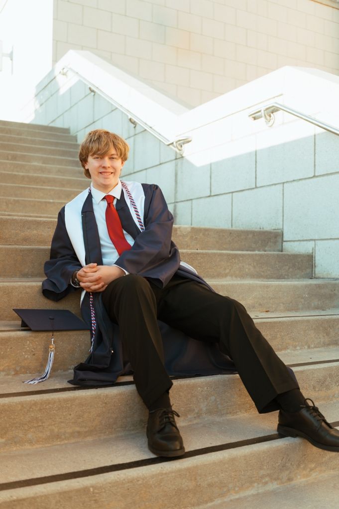 Smiling graduate in cap and gown sits relaxed on outdoor stone steps with diploma.