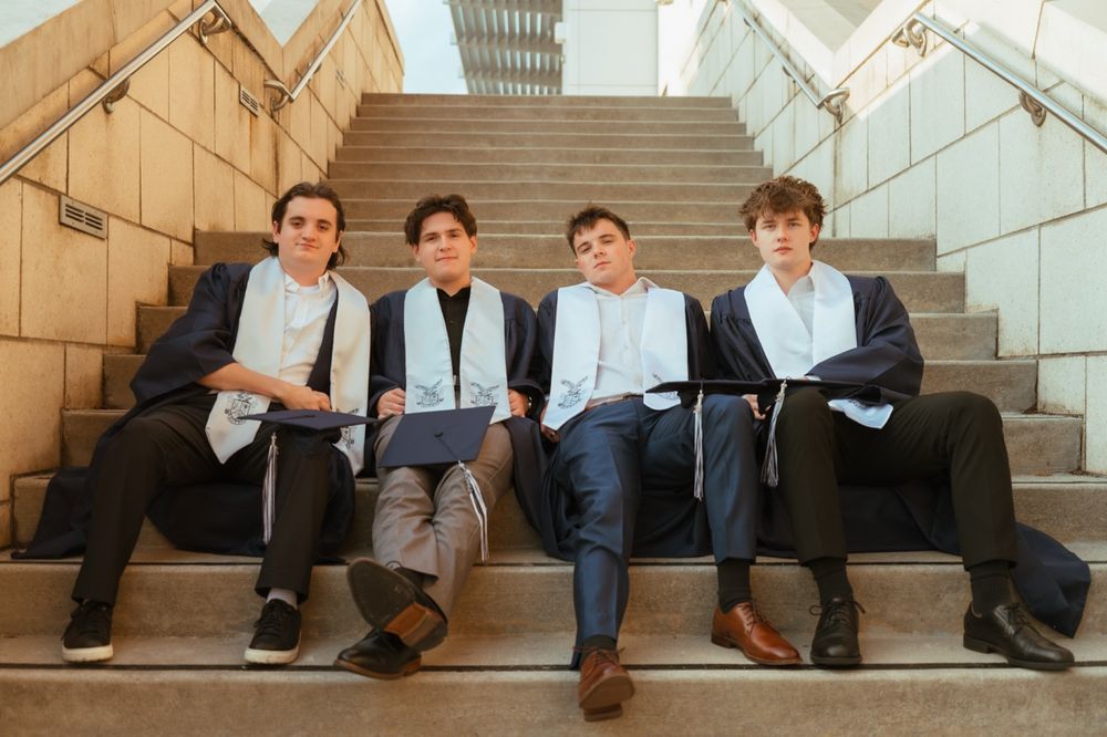 Four young men in suits and graduation stoles sitting casually on outdoor steps.