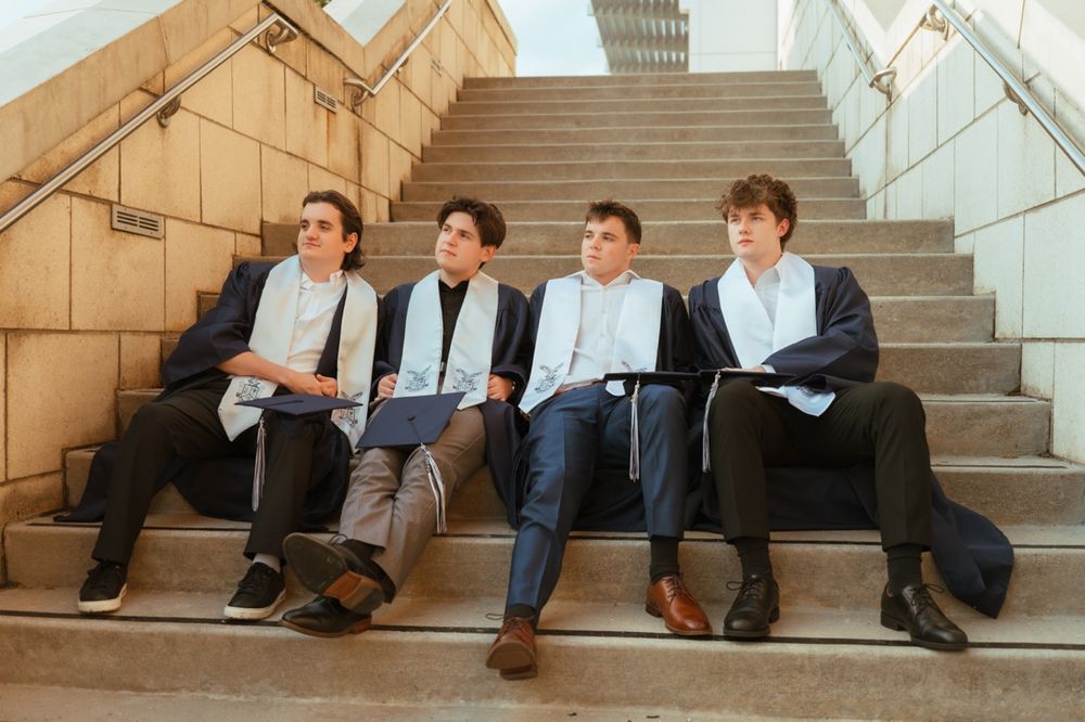 Four male graduates in dark gowns and stoles sit casually on stone staircase steps.