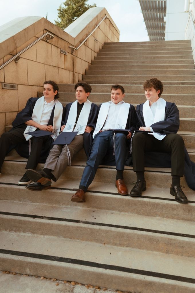 Four young men in graduation gowns and stoles sit relaxed on outdoor concrete steps.