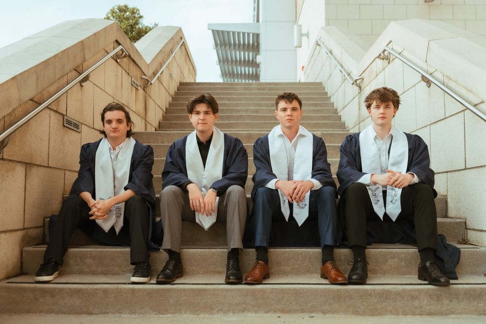 Four young men in graduation attire sitting on outdoor steps, wearing navy suits and white stoles.