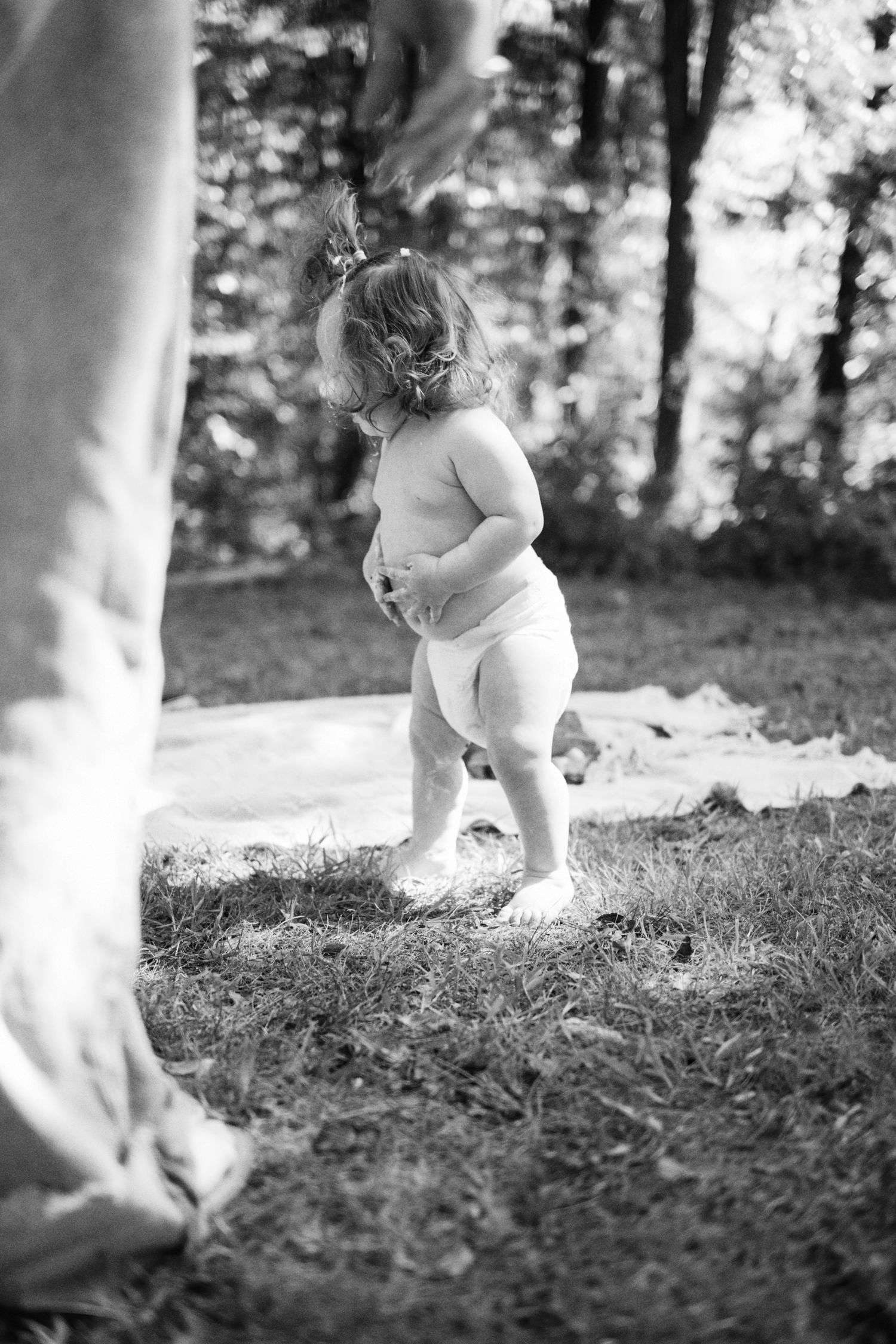 Black and white photo of a toddler in a diaper standing on grass outdoors near trees.