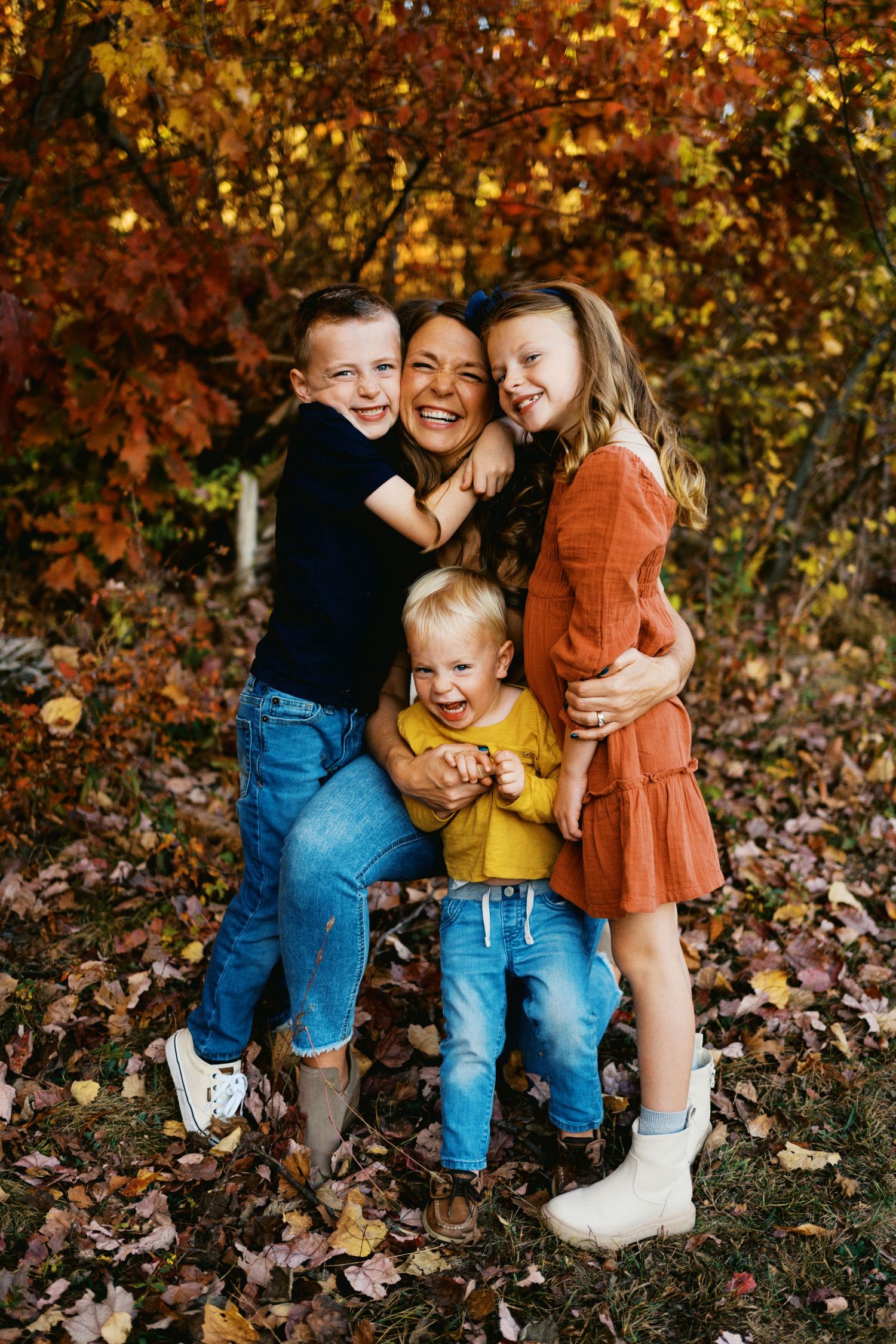 A joyful mother hugs her three children outdoors among colorful autumn leaves and trees in a fall family portrait.
