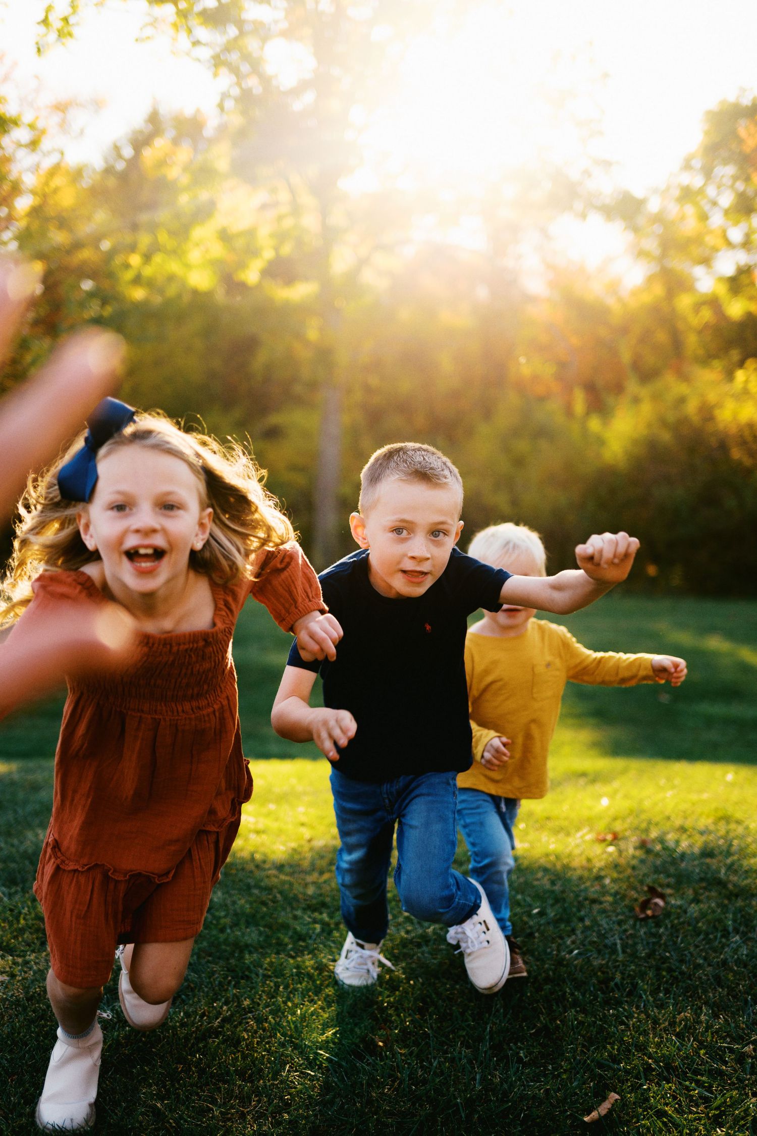 Three joyful children running through a sunlit park, laughing and playing on lush green grass during golden hour.