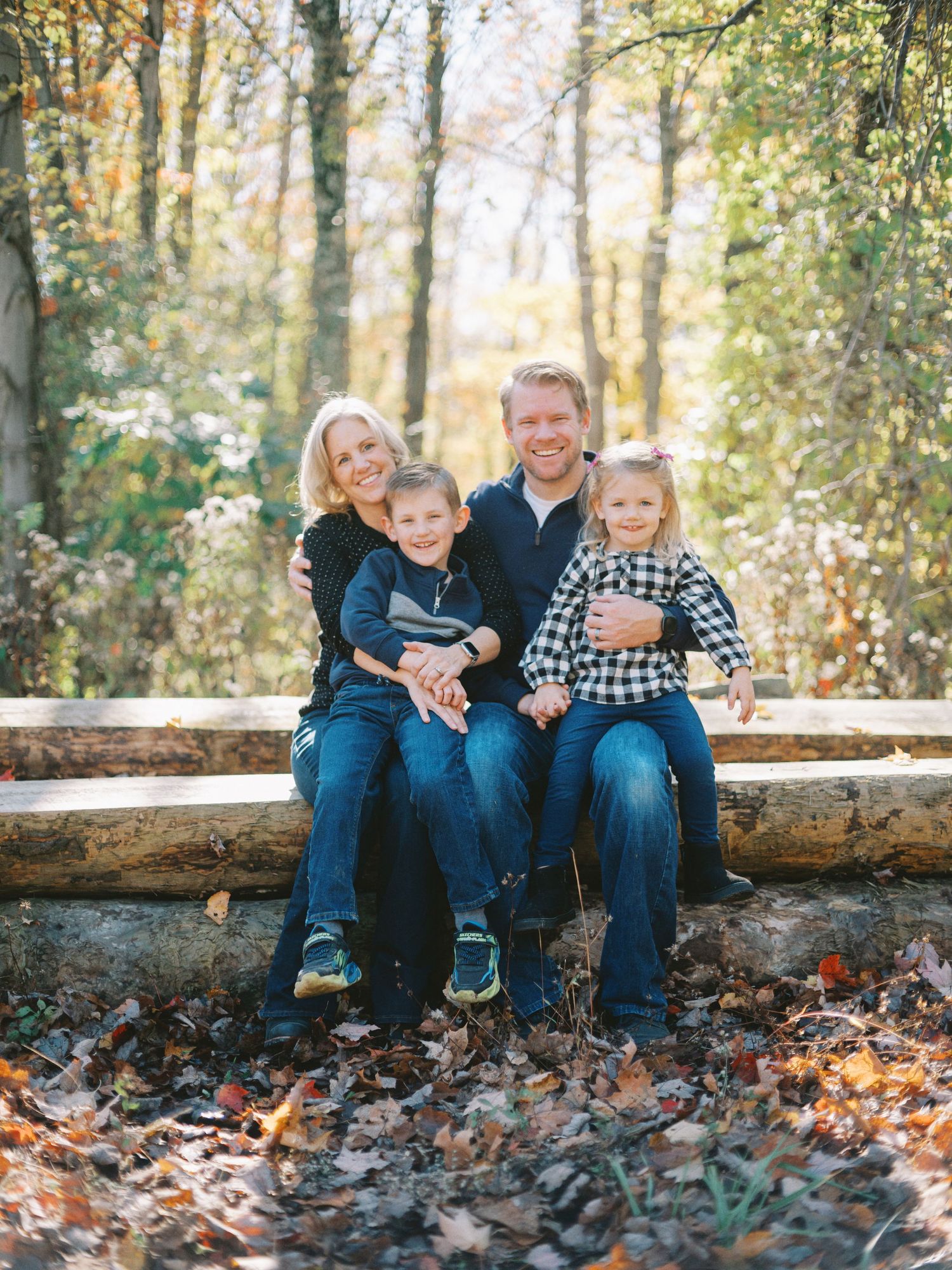 Family of four smiling together on a log in an autumn forest, surrounded by colorful fallen leaves.