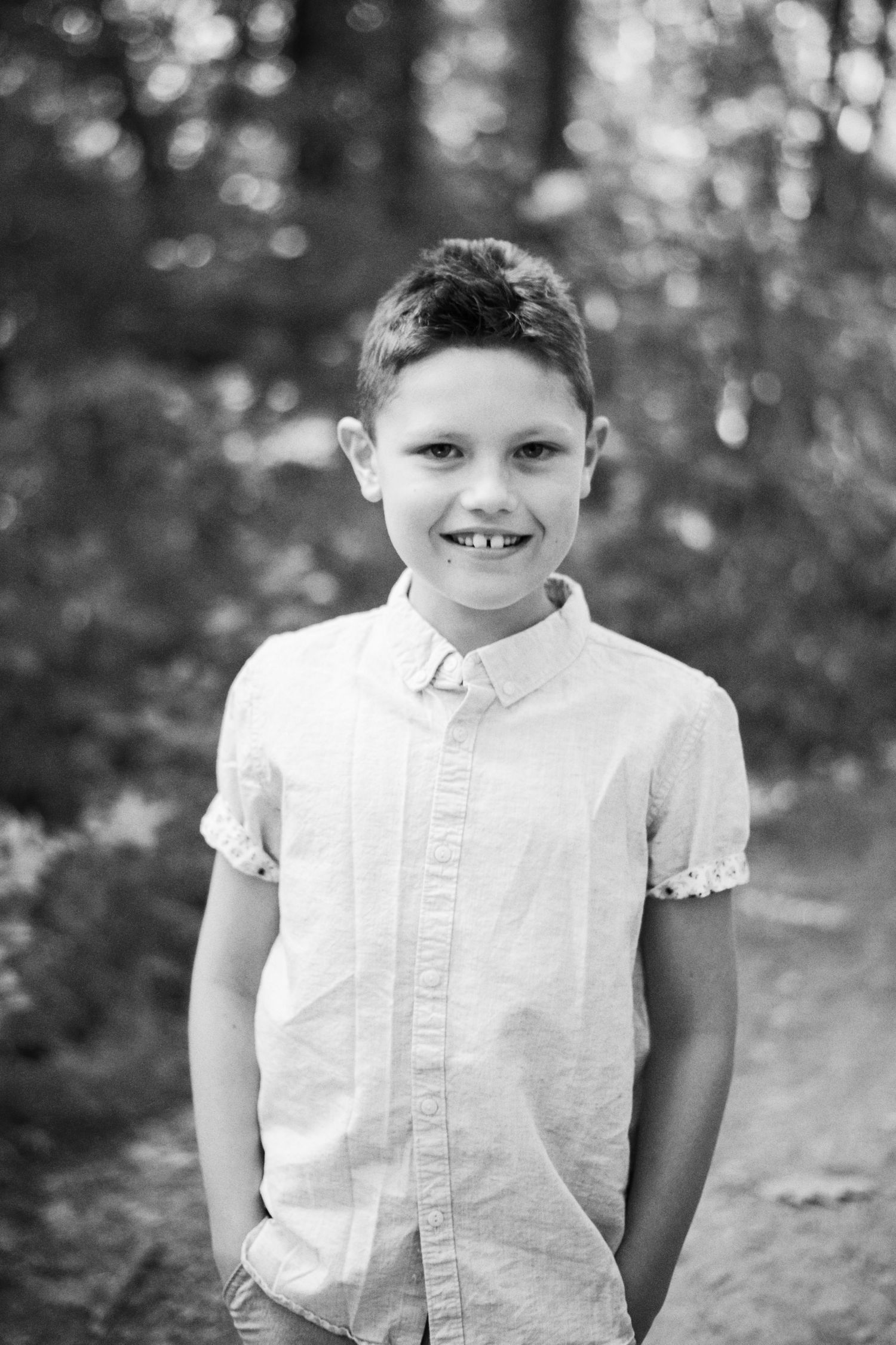 Black and white portrait of a smiling young boy in a button-up shirt standing outdoors in a wooded area.