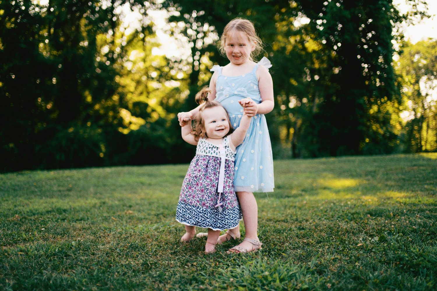 Two young sisters in floral dresses playing together on green grass in a sunny park setting.