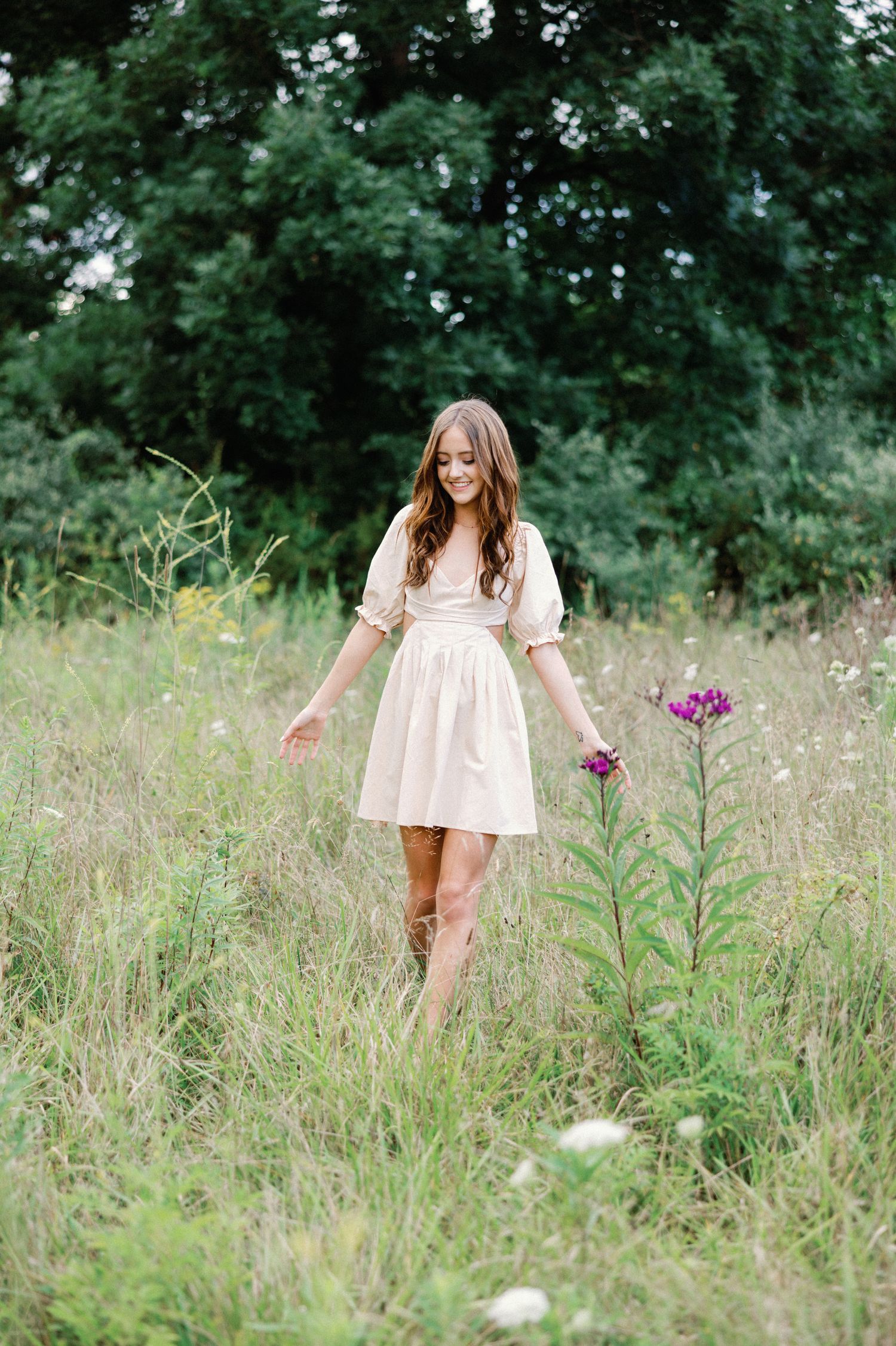 Young woman in a cream dress and cowboy boots walking through a wildflower meadow holding purple flowers.