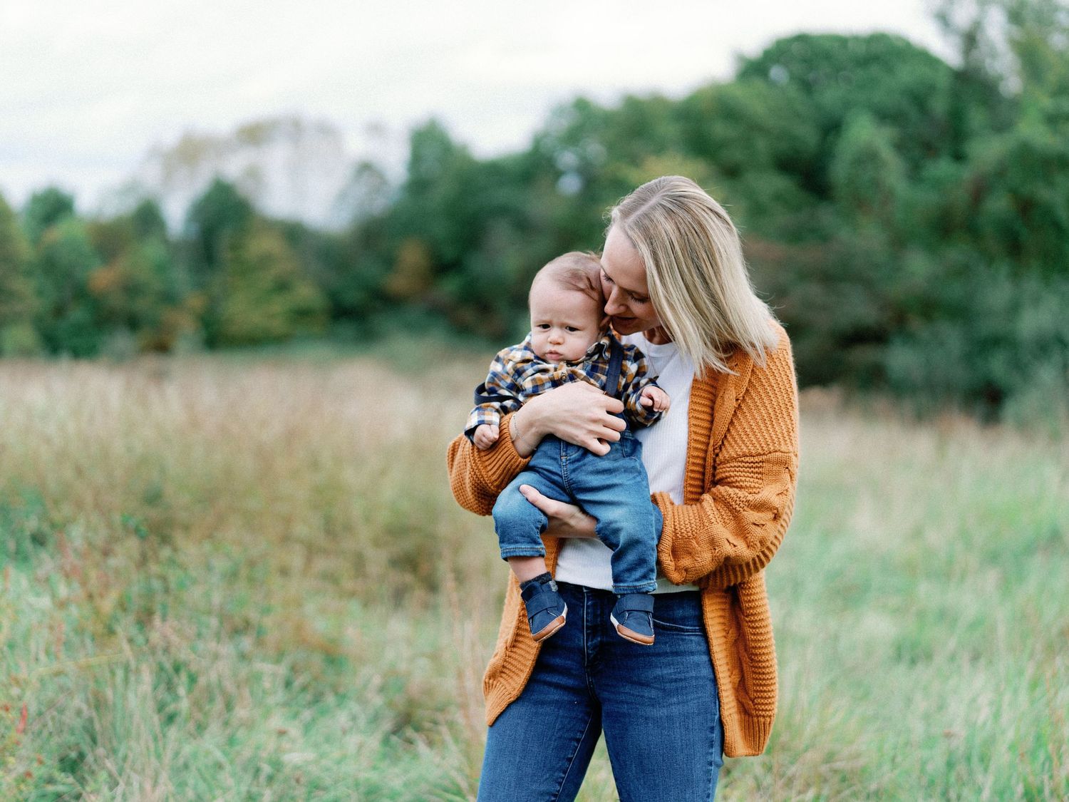 A blonde mother in an orange cardigan cuddles her baby boy in an outdoor fall photo session.