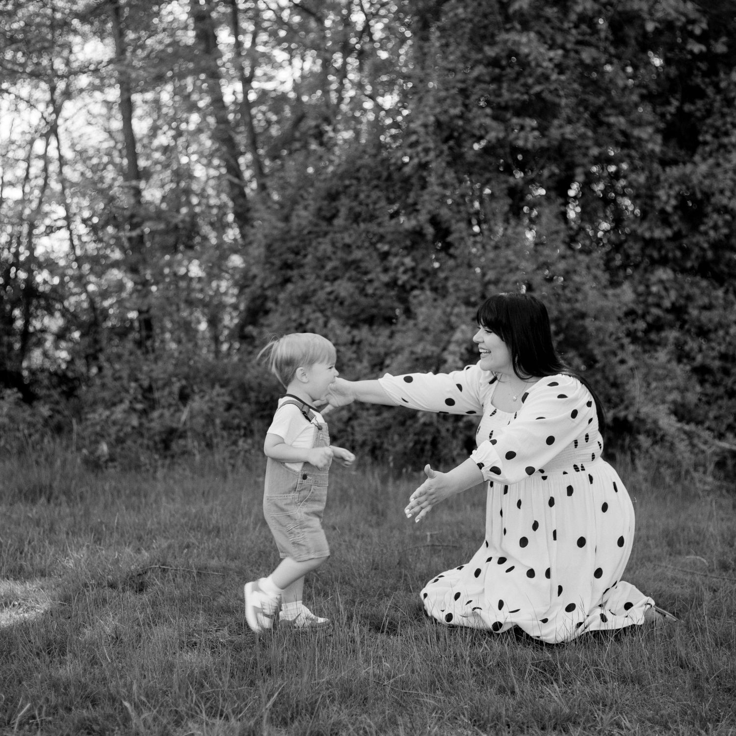 Black and white photo of a mother in polka dot dress kneeling on grass, reaching out to her toddler son outdoors.