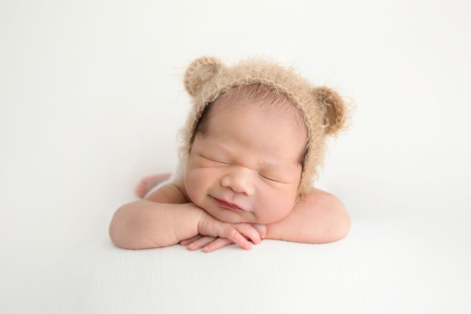 Sleeping newborn baby wearing a fuzzy bear ear bonnet, resting chin on hands against a white background.