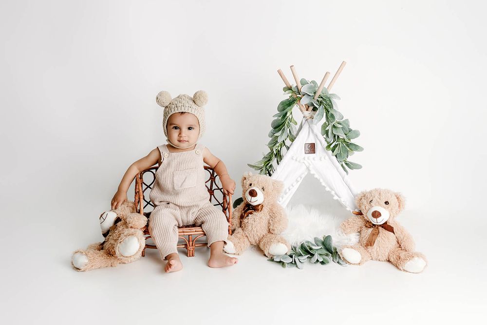 Baby in bear bonnet sits on rattan chair with teddy bears and white teepee backdrop.