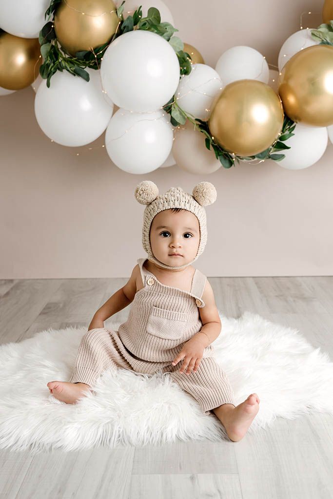 Cute baby in bear bonnet hat and knit romper sits on fur rug with gold and white balloon arch.