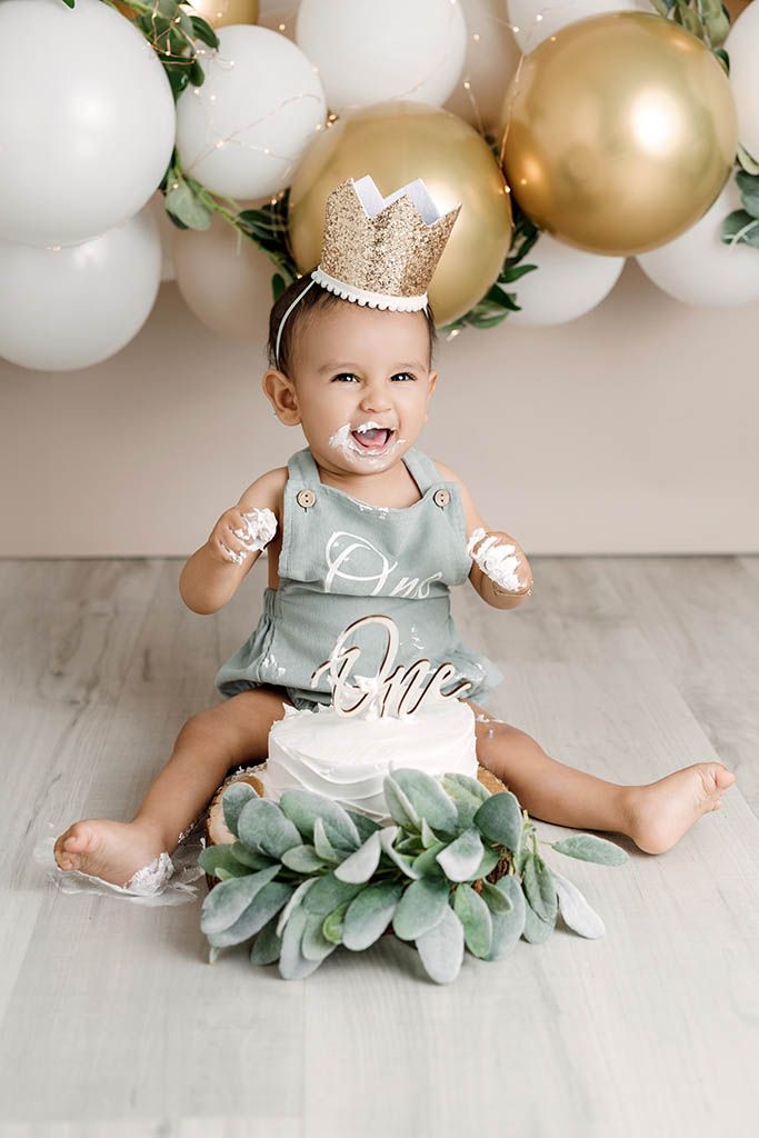 Smiling baby in sage romper and gold crown celebrates first birthday smash cake with white frosting and balloon backdrop.