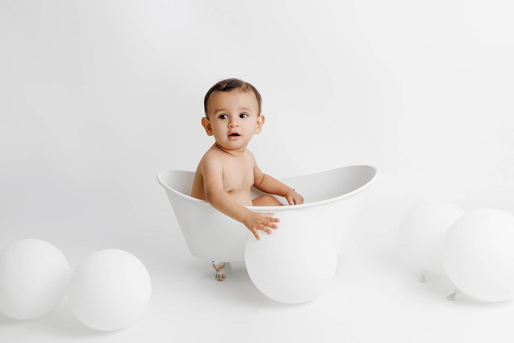 Affordable photographer Menifee photo of cute baby sitting in a small white bathtub surrounded by white balloons on a white background.