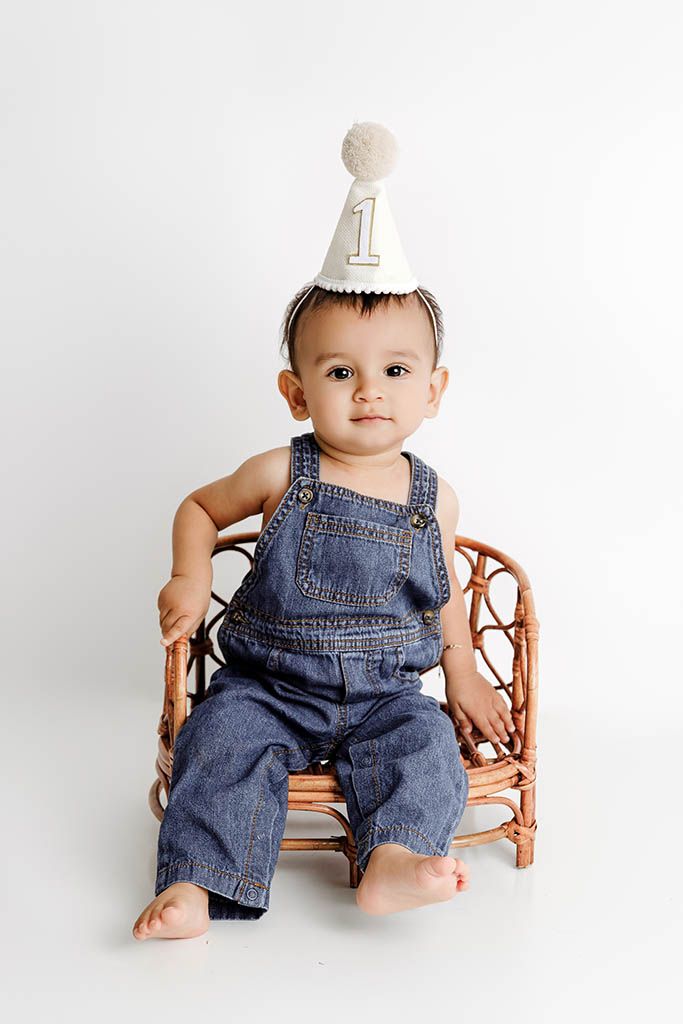 Affordable photographer Menifee photo of baby in denim overalls and number one party hat sits in rattan chair on white backdrop.
