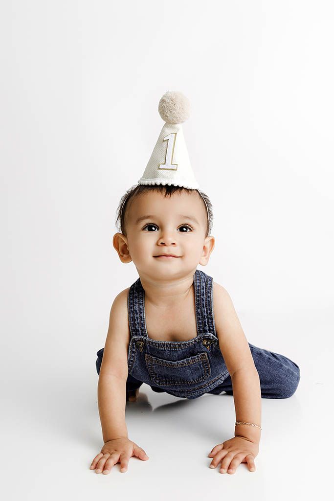 Affordable photographer Menifee photo of cute baby in denim overalls wearing a number 1 birthday party hat, crawling on white background.