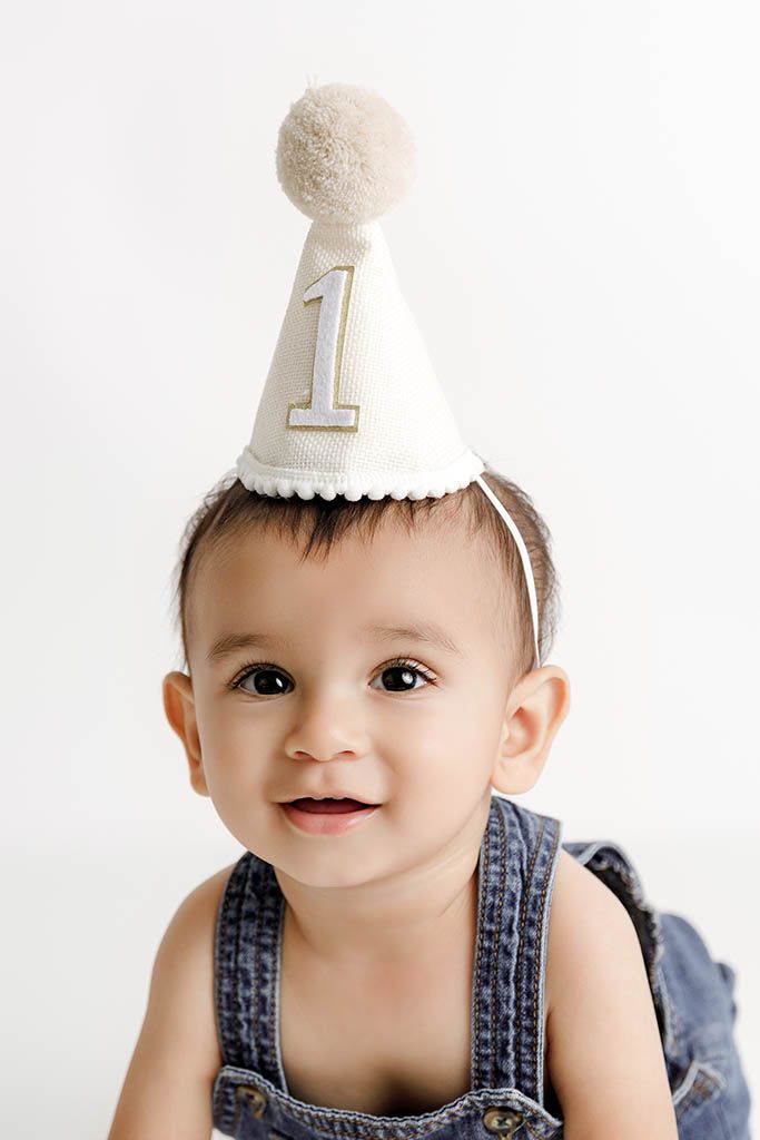 Affordable photographer Menifee photo of adorable baby wearing a number one birthday party hat and denim overalls on a white background.
