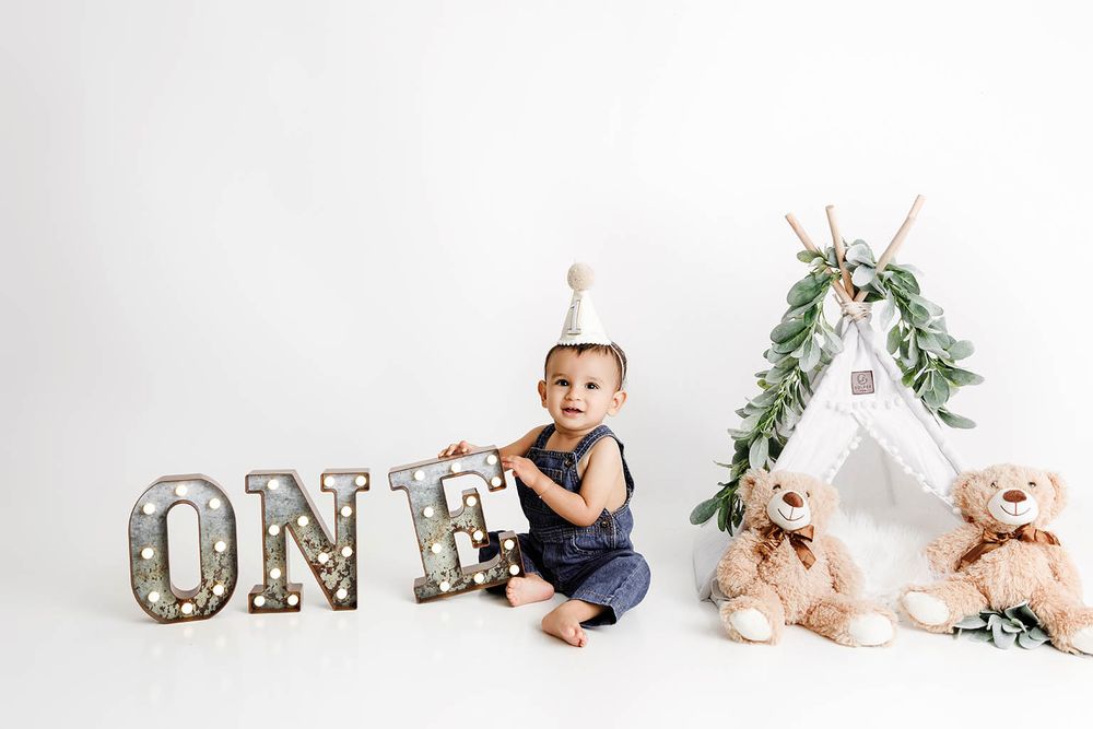 Baby in navy romper poses with ONE marquee letters, teddy bears, and a teepee for first birthday photos.