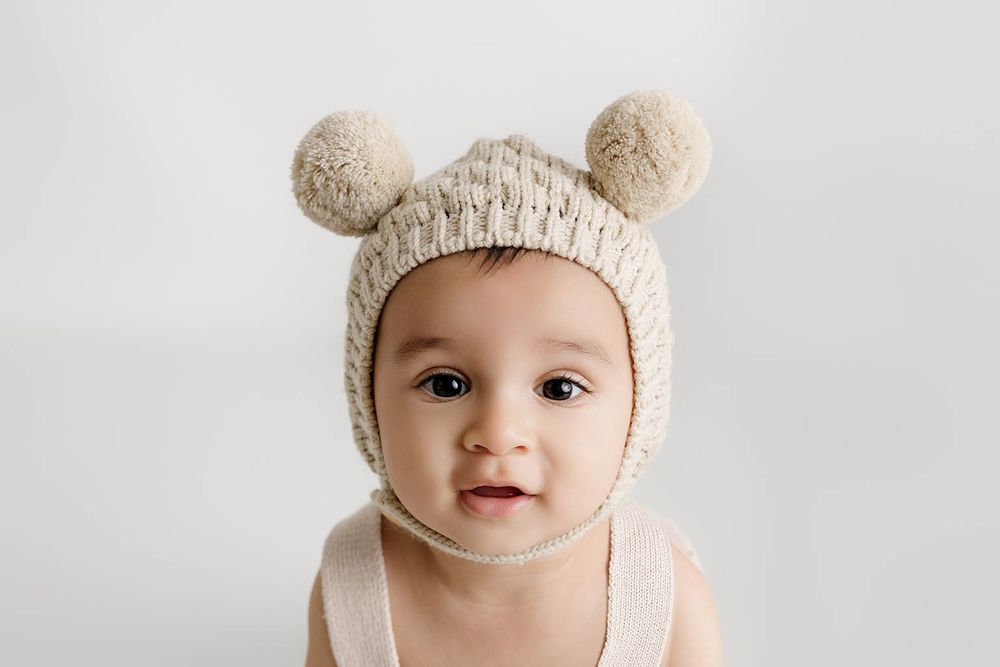 Adorable baby wearing a knitted cream bear ear bonnet hat, looking at camera against white background.